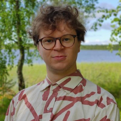 Portrait of Joonas Vaarala. Joonas has light brown, curly hair and glasses. Joonas is wearing a white-collared geometrically patterned shirt.